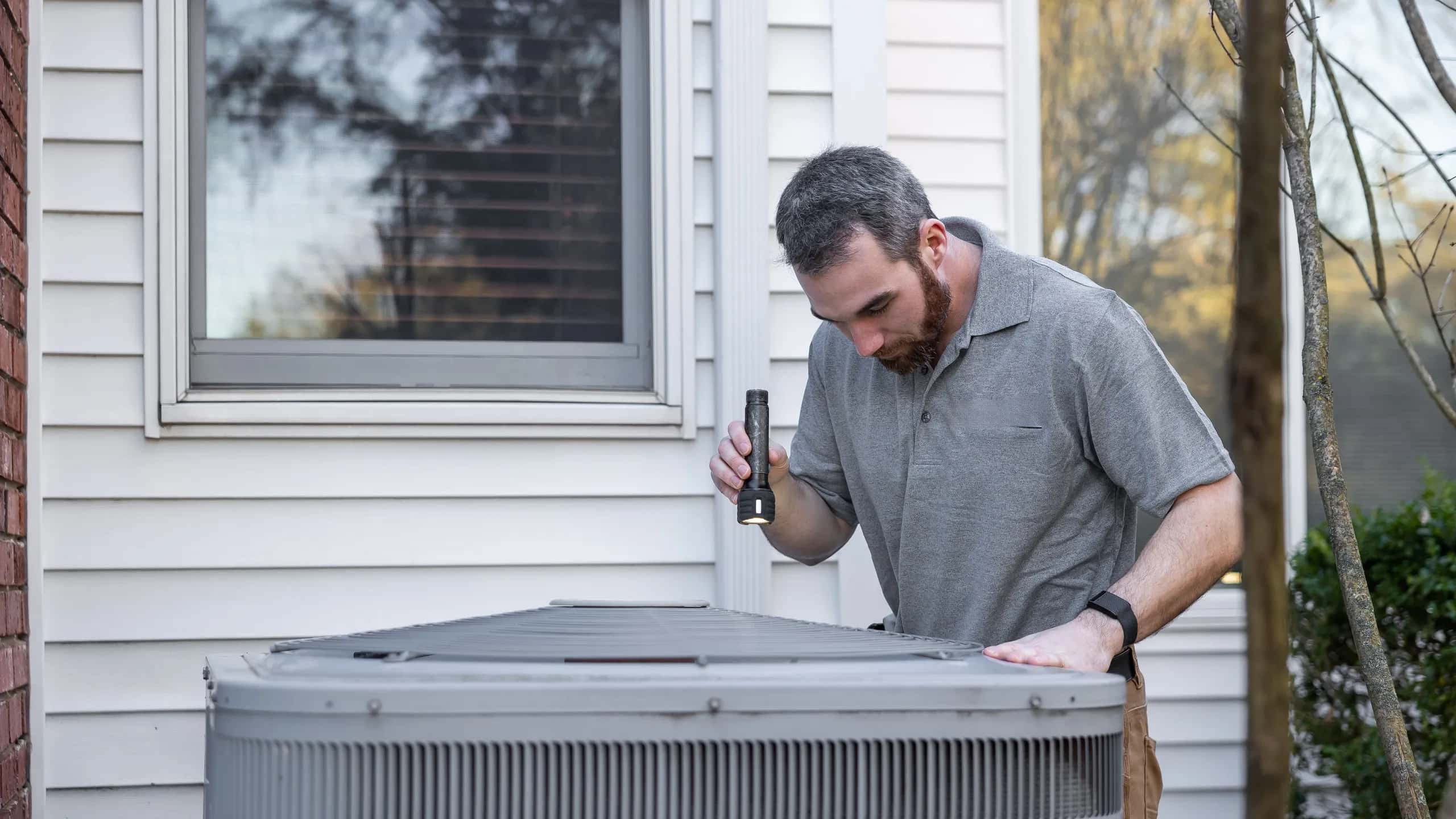 Technician checking an outdoor air conditioning condenser with a flashlight beside a home