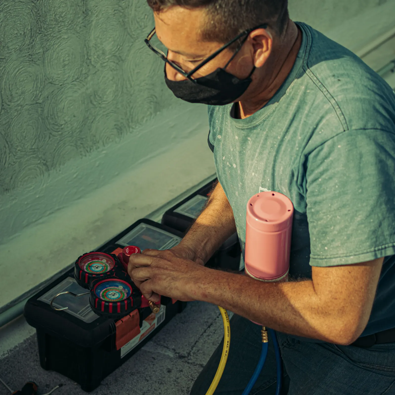 HVAC technician kneeling beside an outdoor unit with refrigerant manifold gauges and an open tool case